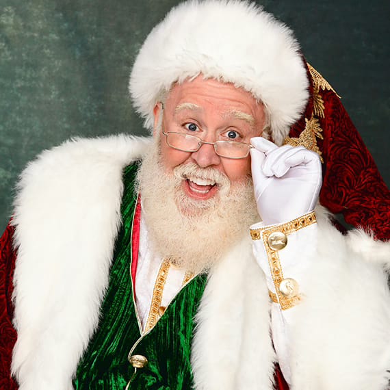 A cheerful Santa Claus in a green vest and fur-trimmed coat, smiling and lifting his glasses.
