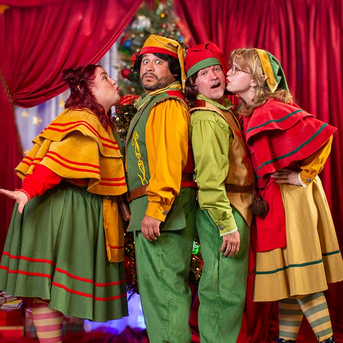 Four House of Kringle elves posing together playfully in front of a Christmas tree.