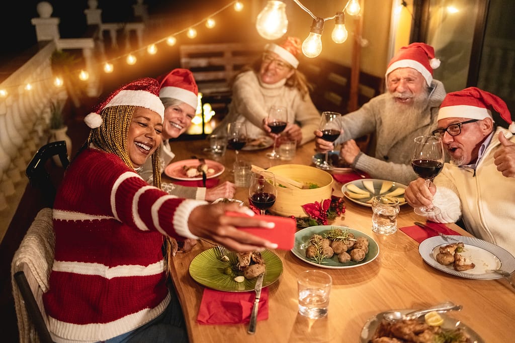 Happy senior friends celebrate Christmas by taking a selfie during dinner at home wearing Santa Claus hats