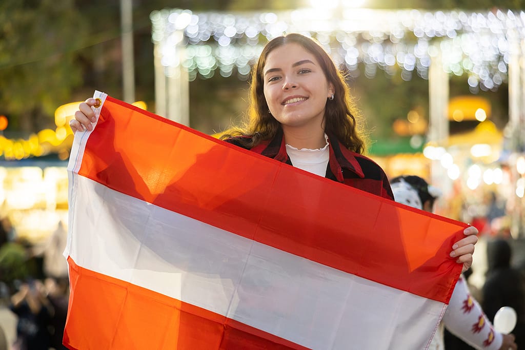 Cheerful young girl having fun at traditional city Christmas market in Austrian city, posing outdoors against blurred background of illuminated shopping stalls with national flag of Austria in hands