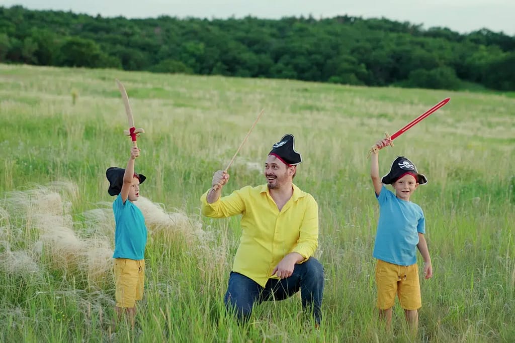 Happy Dad Kids Play Pirates Together On Green Grass. Pirate Father Child With Saber Family Games Outdoors Goes To Treasure Hunt On Pirate Island.