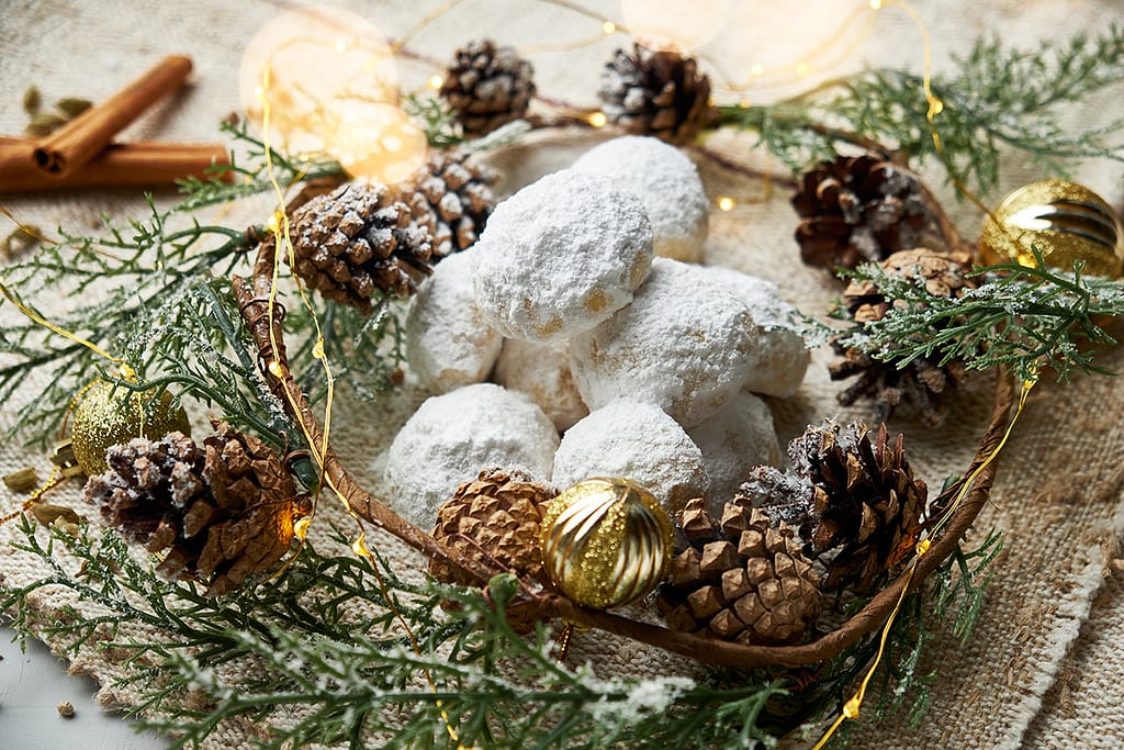 A plate of pfeffernusse surrounded by pine cones, green garland and gold Christmas ornaments.