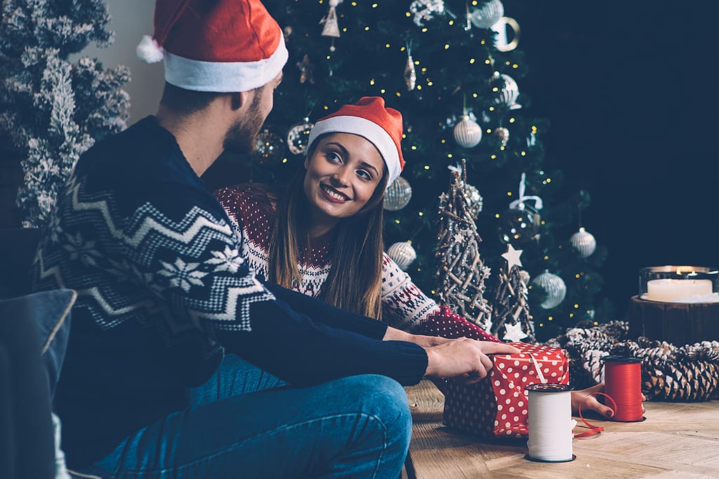 stock photo of a young couple wearing santa hats sitting on a sofa and wrapping gift boxes together box together