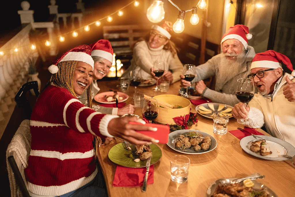 Happy senior friends celebrate Christmas by taking a selfie during dinner at home wearing Santa Claus hats
