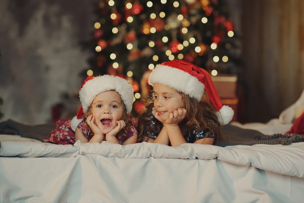 Stock photo of two cute sisters in santa claus hats are sitting on a soft home bed against the background of a christmas tree